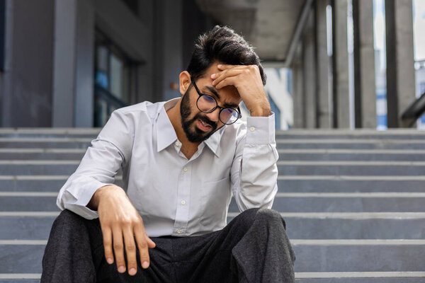 A stressed businessman in despair sits on outdoor steps, expressing worry and frustration, embodying work-related stress.