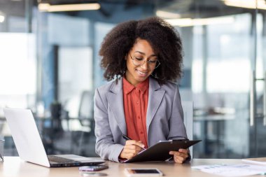 Portatif bilgisayarın yanında otururken pano klasörüne elle notlar yazan mutlu Latin kadın. Üretken işçi işaretlemesi, kişisel etkinliğin izlenmesi için yapılacaklar listesinden tamamlandı.
