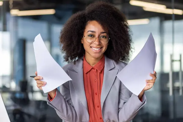 Friendly woman in stylish business suit keeping sheets of paper in both hands on blurred background. Optimistic office receptionist welcoming building visitors with questionnaires to filling.