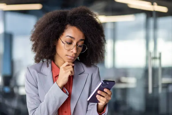 Thoughtful latino woman in grey suit holding chin with hand while looking at smartphone screen indoors. Doubting office worker getting text message with business offer and pondering about answer.