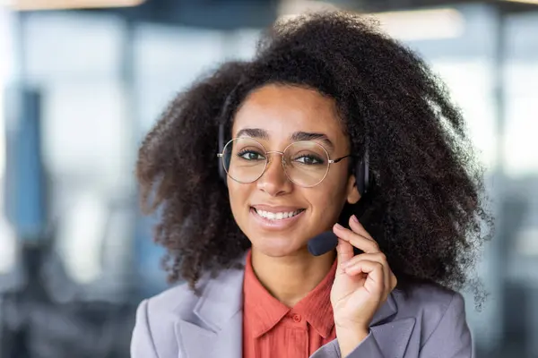 Portrait of black woman with bushy hairstyle fixing microphone on wireless headphones and smiling at camera. Friendly lady in round eyeglasses checking audio accessibility for high quality service.