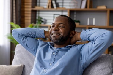 Huzurlu bir ifadeye sahip memnun bir adam kanepede dinlenir, elleri başının arkasında, gözleri kapalı, evinde huzurlu bir anın tadını çıkarır..