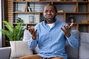 Afro-Amerikalı bir adam omuz silkme ile kafa karışıklığı gösteriyor, iyi dekore edilmiş bir oturma odasında bir kanepede oturuyor..