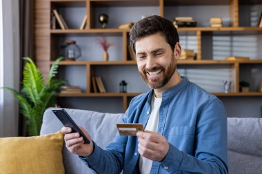 Elinde bir kredi kartı ve akıllı telefon tutan, neşeli bir ifadeyle internetten satın alan sıradan bir erkek..