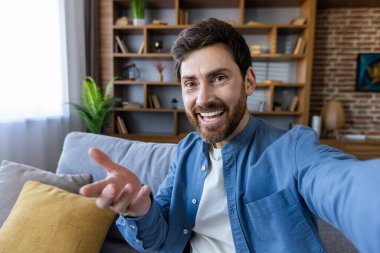 Dostça gülümseyen mutlu bir adam rahat bir oturma odasında selfie çekiyor, neşe ve modern bağlantıyı ifade ediyor..