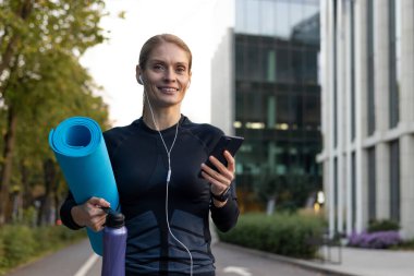 Kendine güvenen kadın profesyonel spor arası, yoga minderi ve su şişesi tutuyor, açık havada telefon kullanıyor..