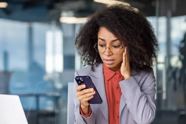 Portrait of worried black woman in eyeglasses rubbing cheek with hand while looking at cellular phone screen. Boss assistant checking online mailbox and noticing big about of unanswered messages.