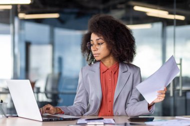 Bir yandan kağıt tutarken bir yandan da taşınabilir bilgisayar klavyesinde yazan resmi kıyafetli sakin bir kadın. Ciddi bir kadın müşterisi için sözleşme yapıyor ve bunu kişisel işyerinde basıyor..