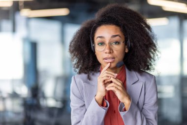 Kablosuz kulaklıklı, parmaklarıyla çeneye dokunan ve kameraya bakan farklı kadın portresi. Odaklanmış yardım hattı operatörü dikkatle müşteri şikayetlerini dinliyor ve çözüm hakkında düşünüyor.