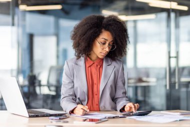 Özenli finans müdürü hesap makinesine güvenmenin doğruluğunu kontrol ediyor ve dizüstü bilgisayarında dizüstü bilgisayarla duran banknotları analiz ediyor. Modern ofisteki şirket giderlerini yöneten takım elbiseli kadın..