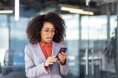 Gri ceketli İspanyol kadın ağzı açık cep telefonu ekranına bakıyor. Yetişkin kadın internet bankacılığı başvurusunu kontrol ediyor ve çalışma yerinde dururken kart dengesi karşısında hayrete düşüyor..