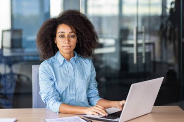 Resmi kıyafetleri olan İspanyol kadın sandalyede oturuyor ve ofis içindeki ahşap masada dizüstü bilgisayarın başında çalışıyor. Özenli sekreter çevrimiçi belgeler hazırlıyor ve patrondan gelen görevleri tamamlamaya hazır..