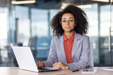 Mikrofonlu kulaklık takan İspanyol kadın ofisin içinde taşınabilir bilgisayarla çalışıyor. Müşteri desteği temsilcisi kameraya bakarken istemci sorununu uzaktan düzeltmeye yardım ediyor.