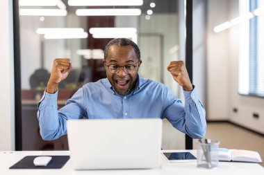 Mavi gömlekli kendinden geçmiş Afro-Amerikan erkek bir ofis ortamında dizüstü bilgisayarıyla çalışırken başarılı bir anı kutluyor..