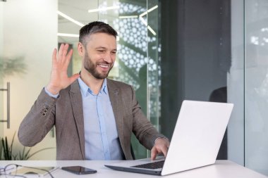 Neşeli iş adamı meslektaşlarını neredeyse dizüstü bilgisayar kullanarak karşılıyor. Modern bir ofiste takım elbise giyiyor, gülümseyerek el sallıyor..
