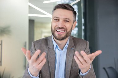 Takım elbiseli gülümseyen profesyonel bir adam parlak çağdaş ofis ortamında dostça bir jest sunuyor, kendine güveni ve ulaşılabilirliği yayıyor..