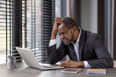 Takım elbiseli bir Afrikalı işadamı modern ofis ortamında dizüstü bilgisayarıyla çalışırken hayal kırıklığına uğramış görünüyor..