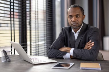 Siyahi takım elbiseli ciddi bir Afro-Amerikan işadamı masasında kollarını kavuşturmuş, modern bir ofis ortamında oturuyor..