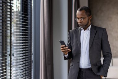 Modern ofis ortamında akıllı telefonunu kullanarak takım elbiseli, olgun Afro-Amerikan iş adamına odaklanmış..