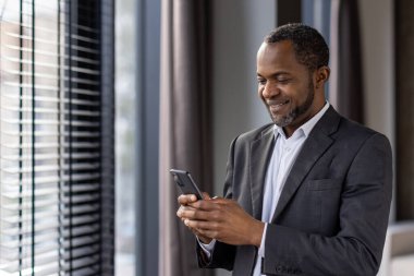 Parlak bir pencerenin yanında akıllı telefonuyla konuşurken gülümseyen resmi takım elbiseli profesyonel Afro-Amerikan erkek..