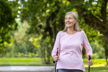 Yeşil, sakin bir parkta İskandinav yürüyüş rutininin keyfini çıkarırken yürüyüş direkleri parlak bir şekilde gülümseyen etkin son sınıf öğrencisi bir kadın..