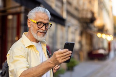 Şık, yaşlı ve gözlüklü bir beyefendi akşam karanlığında şehrin hareketli caddesinde akıllı telefonunu kullanırken gülümsüyor..