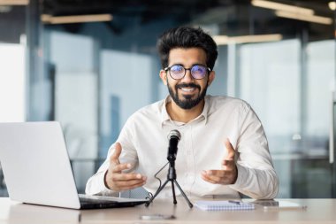 Gülümseyen ve başarılı genç Hintli erkek işadamı mikrofonun önündeki masada oturuyor ve online bir toplantı yapıyor, iş eğitimi alıyor ve kamerayla konuşuyor..