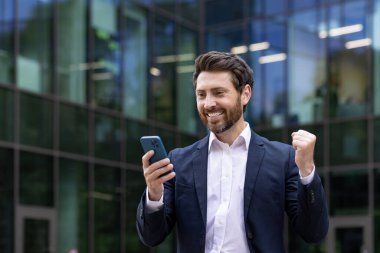 Genç, profesyonel bir işadamı akıllı telefonunu açık havada tutarken yumruğuyla başarılı bir anlaşmayı kutluyor..