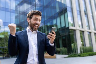 Takım elbiseli neşeli erkek yönetici, cam ofisin dışında akıllı telefonu tutarken muzaffer bir yumruk pompasıyla tezahürat yapıyor..