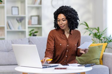 Konsantre ve gülümseyen genç Afrikalı Amerikalı kadın aile bütçesi belgeleri ve dizüstü bilgisayarlarıyla uzaktan çalışıyor, ev hesaplarını ve aile bütçesini kontrol ediyor..