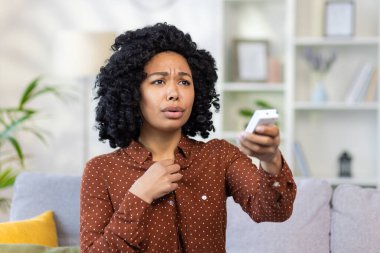 Genç bir Afro-Amerikalı kadının uzaktan kumandayla klimayı açarken çekilmiş yakın plan fotoğrafı. Evdeki kanepede oturuyor ve boğuluyor, iyi hissetmiyor..