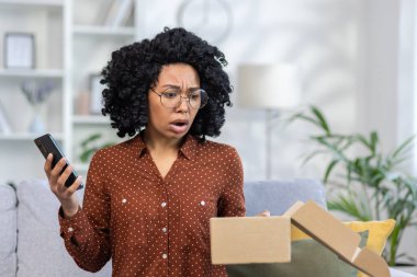 Çevrimiçi paket teslimatı ile ilgili sorunlar. Hüsrana uğramış ve sinirli genç Afrikalı Amerikalı kadın evdeki koltukta oturuyor, elinde telefon ve açık bir kutu var..