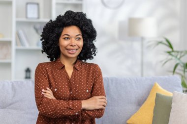 Evdeki kanepede oturan gülümseyen genç bir Afro-Amerikan kadının yakın plan fotoğrafı. Yan tarafa bakıyor, güvenli bir şekilde kollarını göğsünün üzerinden geçiriyor..