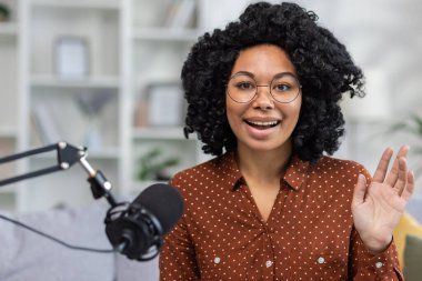 Genç bir Afrikalı Amerikalı kadın çevrimiçi yayın yapıyor, uzaktan eğitim veriyor, ev stüdyosunda sohbet ediyor ve canlı yayına giriyor. Kanepede kameranın önünde oturuyor ve gülümseyerek selamlıyor..