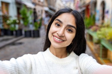 Beyaz kazaklı, gülümseyen bir Hintli kadının yakın plan portresi. Şehir caddesinde selfie çekiyor, kameraya konuşuyor, elinde telefon tutuyor..