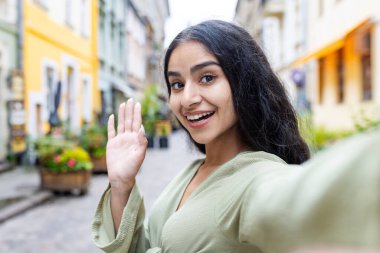 Seyahat eden Hintli genç bir kadının yakın plan portresi, şehir caddesinde yürümek, kendini telefonla çekmek, video görüşmesi yapmak, blog yazmak, el sallamak ve kameraya merhaba demek..