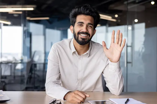 Online business meeting and training. Indian young businessman sitting at the desk in the office, waving to the camera and smiling.