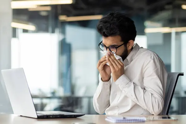 A sick young Indian man is not feeling well at the workplace. He sits at the desk in the office and wipes his nose with a napkin from a runny nose.