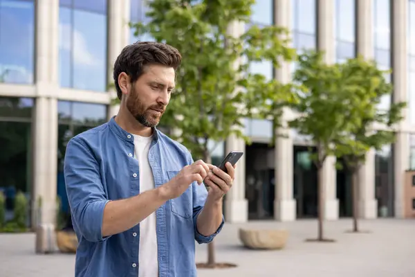 A young man businessman, freelancer stands near an office building and uses a mobile phone.