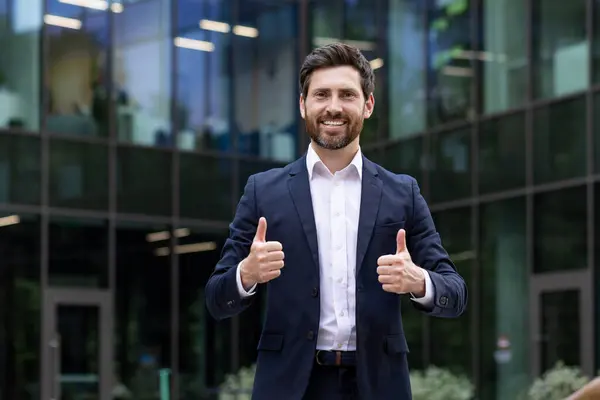 Smiling corporate professional in a suit outside an office building, giving two thumbs up, portraying confidence and success.