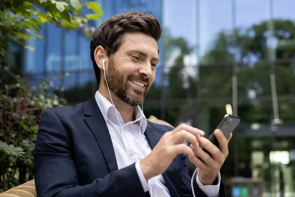 Happy male professional in formal attire outside using mobile phone with earbuds, urban setting in background.