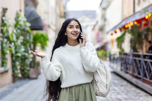 An Indian young smiling woman walks around the city, talks expressively on a mobile phone, has an emotional discussion, gestures with her hands.