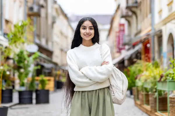 Portrait of a confident Indian young female student in a white sweater standing in the middle of a city street with her arms crossed, on her chest and looking at the camera with a smile.