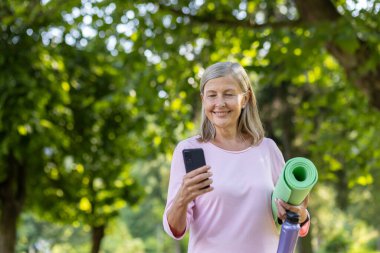 Kolunun altında yoga minderi olan gülümseyen yaşlı bir kadın güneşli bir parkta telefonunu kontrol ediyor. Sağlık ve rahatlama yayıyor..