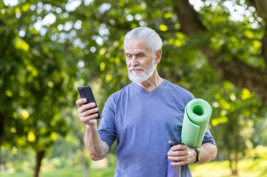 Yeşil yoga minderi tutan yaşlı bir adam dışarıda telefonunu kontrol ediyor, sağlık ve teknolojiyi somutlaştırıyor..