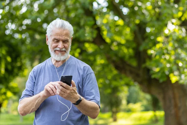 Joyful older man with headphones selects tunes on his phone amidst lush greenery. Representing active seniors and technology.