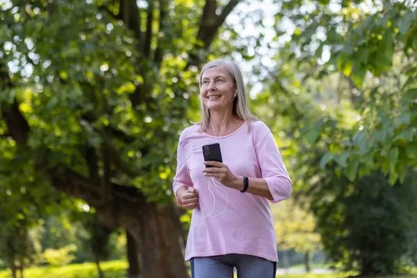 An elderly woman with grey hair smiles as she listens to music and jogs in a vibrant green park.