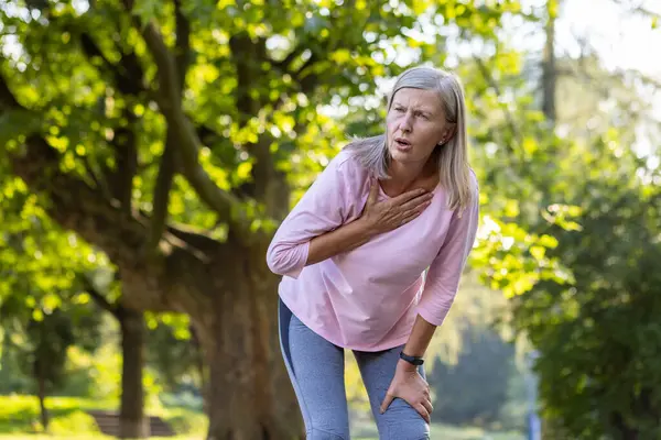 An elderly woman pauses her workout in the park, looking distressed with a hand over her chest, possibly indicating chest pain.