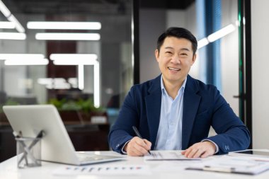 Takım elbiseli gülümseyen Asyalı genç bir adamın portresi ofiste dizüstü bilgisayarla oturuyor ve kameraya bakıyor, belgeler ve verilerle çalışıyor..