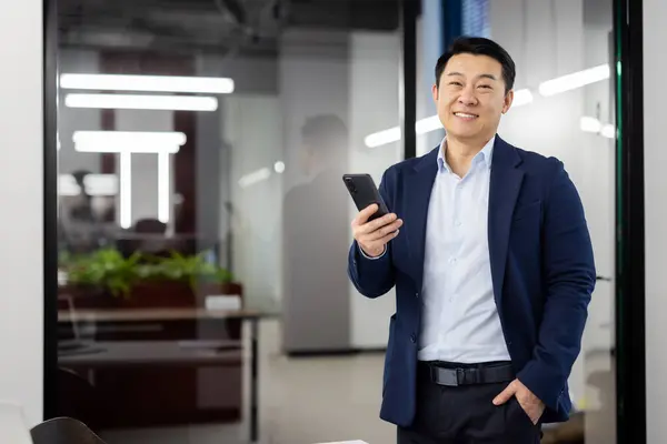 Portrait of successful young Asian man in suit standing in office, holding hand in pocket and mobile phone in other hand, looking confidently and smiling at camera.